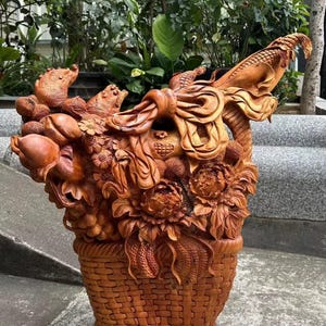 Puede incluir: Una detallada escultura de madera de una cesta rebosante de frutas y flores. La cesta está intrincadamente tallada con una textura tejida. La escultura presenta melocotones, peonías y otros elementos florales, todos en tonos marrones.