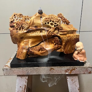 Könnte beinhalten: Ein hölzerner Räuchergefäß in Form einer Berglandschaft mit einem kleinen Haus, Bäumen und einem Wasserfall. Rechts sitzt eine kleine, lächelnde Mönchsfigur. Der Brenner steht auf einem schwarzen Sockel.