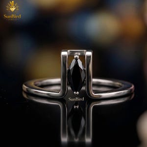 Puede incluir: Un anillo de plata con una gema negra de corte marquesa engastada en un diseño abierto y moderno. El anillo tiene un acabado pulido y la marca "SunBird Jewels" es visible.
