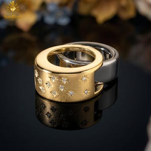 Könnte beinhalten: Zwei Ringe, einer goldfarben und einer silberfarben, werden gezeigt. Der goldfarbene Ring hat eingearbeitete sternförmige Designs mit kleinen, funkelnden Steinen. Der silberfarbene Ring hat eine glatte, polierte Oberfläche. Die Ringe sind von SunBird Jewels.