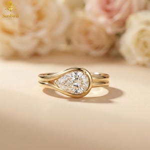 Può includere: Un anello di fidanzamento in oro con un diamante a forma di pera. L'anello ha una fascia delicata e un'incastonatura a castone che mette in risalto la brillantezza del diamante. Lo sfondo è una composizione floreale morbida e sfocata.