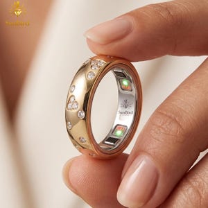 May include: A gold smart ring featuring a polished exterior with embedded round clear stones. The inner band is silver-toned with the text SunBird Jewels and two small rectangular sensors displaying glowing green and red indicator lights.
