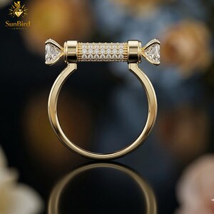 Puede incluir: Un anillo de oro con un dise&ntilde;o &uacute;nico. El anillo presenta una barra central incrustada con peque&ntilde;as piedras claras, flanqueada por piedras claras m&aacute;s grandes, talladas en forma de tri&aacute;ngulo. La banda del anillo es de oro pulido. El logotipo de SunBird Jewels es visible.