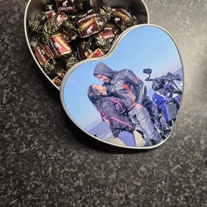 Op de afbeelding: Een hartvormige blikken doos gevuld met individueel verpakte chocolaatjes. Het deksel toont een foto van een stel in motorkleding, dat elkaar omhelst bij een motorfiets. De doos is open en toont de chocolaatjes.