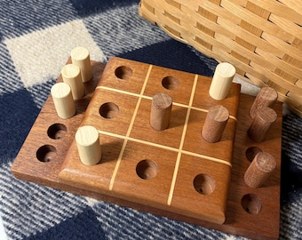 以下が含まれることがあります： 明るい茶色と濃い茶色のペグが付いた木製の三目並べゲーム。ゲーム盤は、明るい木材のアクセントが付いた濃い茶色の木材で作られています。ゲームは青と白のチェック柄の毛布の上に置かれ、背景には柳のバスケットがあります。