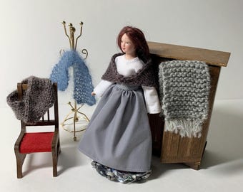 May include: A miniature doll dressed in a gray skirt, white top, and brown shawl stands near a wooden cabinet. A small red chair and a brass coat rack with blue and brown scarves are also present. The scene evokes a cozy, handcrafted aesthetic.