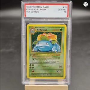 Könnte beinhalten: Eine bewertete 1999 Pokémon Game Venusaur - Holo 1st Edition Karte. Die Karte zeigt eine blaue und grüne Venusaur-Illustration mit einer großen orangefarbenen Blume. Die Karte ist in einem transparenten Schutzgehäuse eingeschlossen.