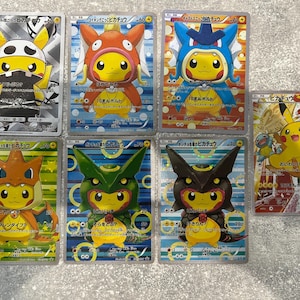 May include: A collection of seven rectangular trading cards featuring the yellow character Pikachu wearing various costumes, including a skull mask, fish, dragon, and other creature-themed outfits with Japanese text printed on each card.