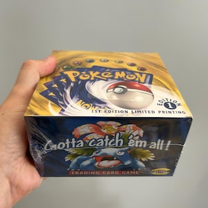 Pode incluir: Uma caixa de boosters do jogo de cartas colecionáveis Pokémon selada com topo amarelo, gráfico de Poké Bola e o texto 1st Edition Limited Printing. O painel frontal azul exibe o personagem Venusaur, o texto Gotta catch 'em all! e o logo.