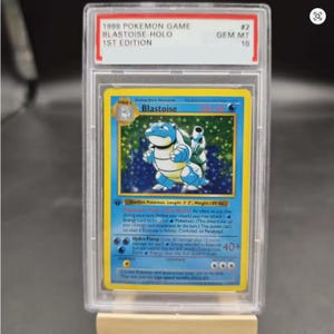 Könnte beinhalten: Eine bewertete 1999 Pokémon Game Blastoise Holo 1st Edition Karte. Die Karte zeigt eine blaue Blastoise-Illustration mit gelbem Rand. Die Karte ist in einem transparenten Schutzgehäuse mit der Note GEM MT 10.