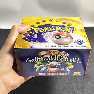 Puede incluir: Una caja sellada del juego de cartas coleccionables Pokémon. La caja es amarilla y azul con el logo de Pokémon y una Pokéball. El texto en la caja dice "Gotta catch 'em all!" y "1st Edition Limited Printing".
