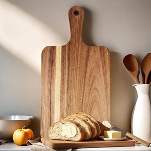 May include: A large wooden cutting board with a handle, bread slices with butter, and a small pumpkin. The cutting board is made of light and dark brown wood. A white vase holds wooden spoons.
