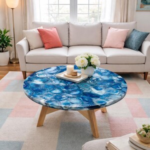 May include: A circular coffee table featuring a blue agate stone top and light wood legs. The table is positioned in front of a cream sofa with pink and blue cushions. A white rug is beneath the table, in front of a window.