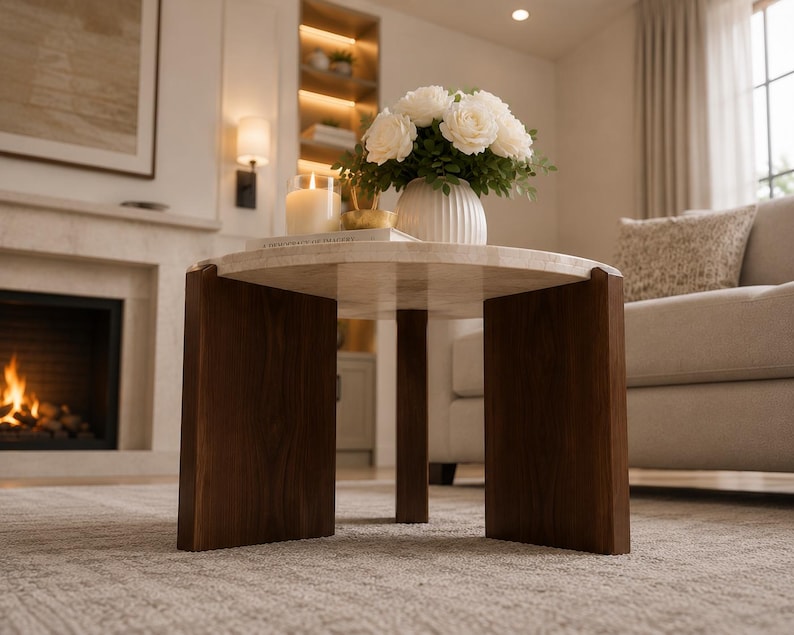 Puede incluir: Una mesa de centro redonda con una superficie de piedra de color claro con un acabado liso y pulido. La mesa se apoya sobre tres patas gruesas de madera de color marr&oacute;n oscuro dispuestas en tr&iacute;pode, proporcionando una base firme.
