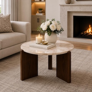 Puede incluir: Una mesa de centro redonda con una superficie de m&aacute;rmol beige pulido con vetas naturales y tres patas de madera de color marr&oacute;n oscuro. Sobre la mesa hay un jarr&oacute;n blanco acanalado con rosas blancas, una vela encendida y un peque&ntilde;o cuenco dorado.