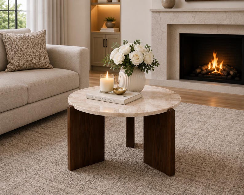 Puede incluir: Una mesa de centro redonda con una superficie de m&aacute;rmol beige pulido con vetas naturales y tres patas de madera de color marr&oacute;n oscuro. Sobre la mesa hay un jarr&oacute;n blanco acanalado con rosas blancas, una vela encendida y un peque&ntilde;o cuenco dorado.