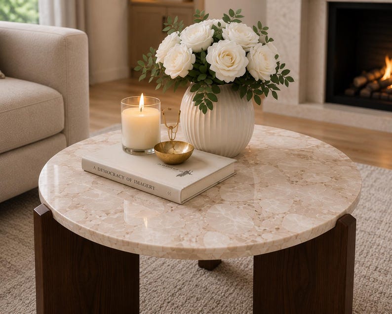 Puede incluir: Una mesa de centro redonda con una superficie de piedra beige y patas de madera marr&oacute;n oscuro. Sobre la mesa hay un jarr&oacute;n blanco acanalado con rosas blancas, una vela encendida en un vaso, un cuenco dorado y un libro titulado A Democracy of Imagery.