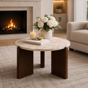 Puede incluir: Una mesa de centro redonda con una superficie de m&aacute;rmol beige claro con un sutil patr&oacute;n veteado y tres patas de madera marr&oacute;n oscuro. Sobre la mesa hay un jarr&oacute;n blanco acanalado con flores blancas, una vela encendida y un libro blanco.