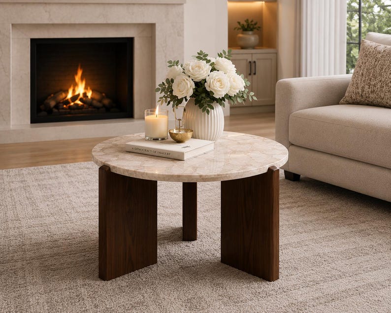 Puede incluir: Una mesa de centro redonda con una superficie de m&aacute;rmol beige claro con un sutil patr&oacute;n veteado y tres patas de madera marr&oacute;n oscuro. Sobre la mesa hay un jarr&oacute;n blanco acanalado con flores blancas, una vela encendida y un libro blanco.