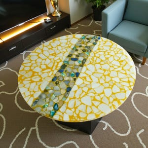 May include: A round coffee table with a yellow and white mosaic top. A diagonal stripe of blue, green, and gray stones runs across the center. The table is supported by a black base.