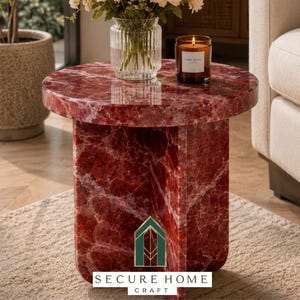 May include: A round side table crafted from deep red marble with prominent white veining. The table features a cylindrical top supported by a cross-shaped base. A clear glass vase with flowers and a brown candle sit on the polished marble surface.