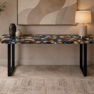 May include: A rectangular console table featuring a tabletop composed of polished blue, brown, and white agate slices. The table is supported by two black metal U-shaped legs. The surface has a glossy finish that reflects light across the stone pattern.
