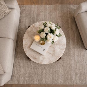 Puede incluir: Una mesa de centro redonda con una superficie de piedra texturizada en color beige claro. Sobre la mesa hay un libro blanco titulado A Democracy of Imagery, un cuenco dorado peque&ntilde;o, una vela encendida y un ramo de rosas blancas.