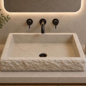 May include: A rectangular stone vessel sink with a rough, chiseled exterior texture and a smooth interior basin. The sink features a central drain hole and is paired with a matte black wall-mounted faucet and two matching circular control handles.