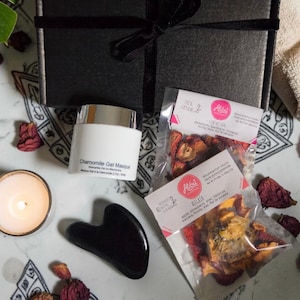 May include: A black gift box with a black ribbon, a white jar labeled Chamomile Gel Masque, two clear pouches of dried fruit labeled Alibú, a black heart-shaped gua sha tool, and a small lit candle on a white surface with scattered dried petals.