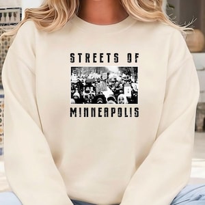 May include: Cream-colored sweatshirt with the text "STREETS OF MINNEAPOLIS" in black block letters. The graphic in the center is a black and white image of a crowd of people. The sweatshirt is a casual style.
