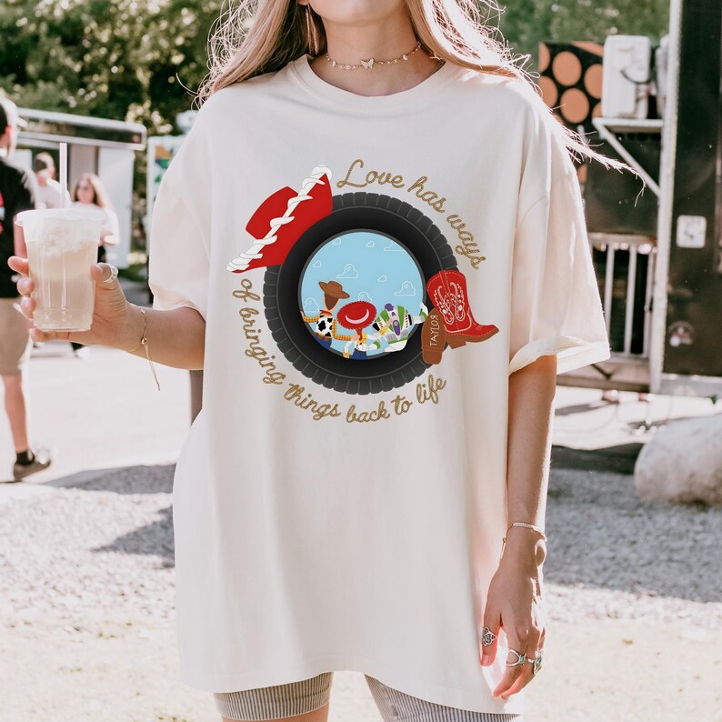 K&ouml;nnte beinhalten: Ein cremefarbenes T-Shirt mit einer Grafik eines schwarzen Reifens, der eine blaue Himmelsszene mit Zeichentrickfiguren umgibt. Ein roter Cowboyhut und ein roter Cowboystiefel befinden sich auf dem Reifen. Der Text lautet, Love has ways of bringing things back to life.