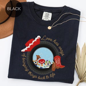 K&ouml;nnte beinhalten: Ein schwarzes T-Shirt mit Rundhalsausschnitt und einem Grafikdruck, der einen Reifen zeigt, welcher eine Szene mit drei Zeichentrickfiguren, einem roten Cowboyhut und einem roten Cowboystiefel umrahmt. Der Text lautet Love has ways of bringing things back to life.