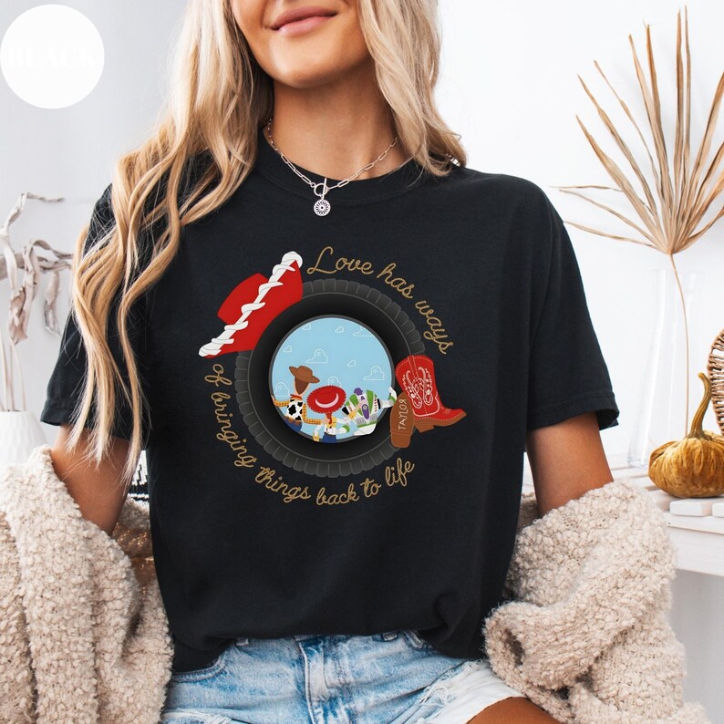 K&ouml;nnte beinhalten: Ein schwarzes T-Shirt mit einer Grafik, die einen Reifen zeigt, der eine Szene mit Zeichentrickfiguren, einen roten Cowboyhut und einen roten Cowboystiefel umrahmt. Der Text "Love has ways of bringing things back to life" steht in goldener Schrift.