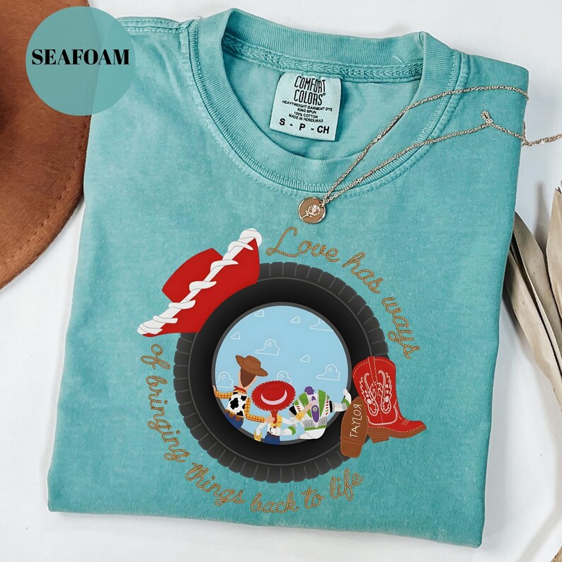K&ouml;nnte beinhalten: Ein meergr&uuml;nes Kurzarm-T-Shirt mit einer Grafik aus einem schwarzen Reifen, der Zeichentrickfiguren, einen roten Cowboyhut und einen roten Cowboystiefel umrahmt. Der Text lautet: Love has ways of bringing things back to life, in Gold.