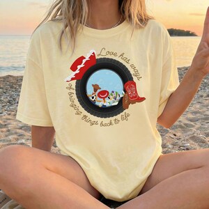 K&ouml;nnte beinhalten: Ein blassgelbes T-Shirt mit einer Grafik eines schwarzen Reifens, der eine blaue Himmelsszene mit Zeichentrickfiguren einrahmt. Ein roter Cowboyhut und ein roter Cowboystiefel liegen auf dem Reifen. Der Text lautet, Love has ways of bringing things back to life.