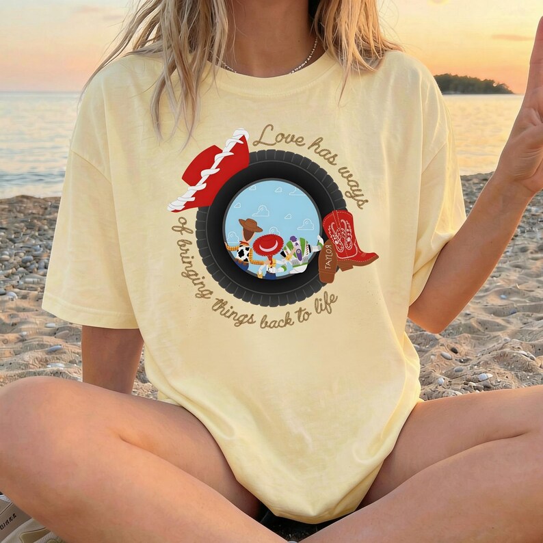 K&ouml;nnte beinhalten: Ein blassgelbes T-Shirt mit einer Grafik eines schwarzen Reifens, der eine blaue Himmelsszene mit Zeichentrickfiguren einrahmt. Ein roter Cowboyhut und ein roter Cowboystiefel liegen auf dem Reifen. Der Text lautet, Love has ways of bringing things back to life.