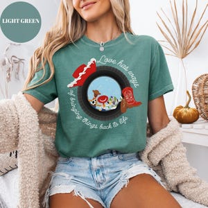 K&ouml;nnte beinhalten: Ein hellgr&uuml;nes Kurzarm-T-Shirt mit einer Grafik eines schwarzen Reifens, der eine Szene mit einem Cowboyhut, einem roten Stiefel und Zeichentrickfiguren einrahmt. Der Text "Love has ways of bringing things back to life" ist in wei&szlig;er Schreibschrift aufgedruckt.