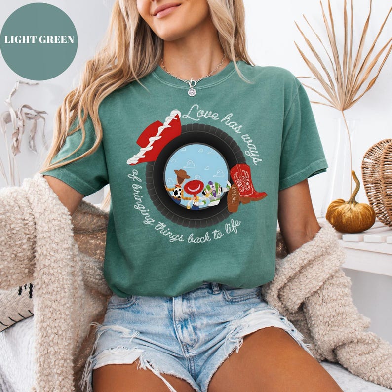 K&ouml;nnte beinhalten: Ein hellgr&uuml;nes Kurzarm-T-Shirt mit einer Grafik eines schwarzen Reifens, der eine Szene mit einem Cowboyhut, einem roten Stiefel und Zeichentrickfiguren einrahmt. Der Text "Love has ways of bringing things back to life" ist in wei&szlig;er Schreibschrift aufgedruckt.