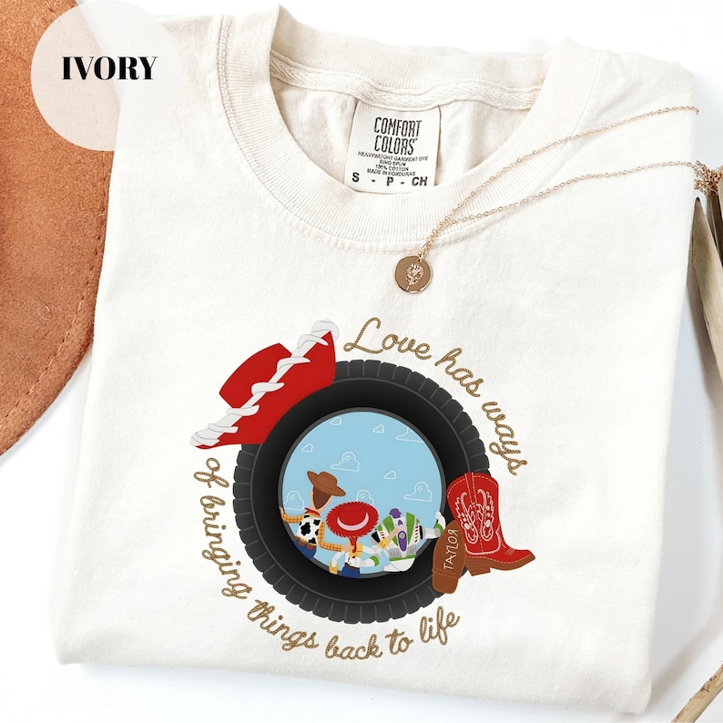 K&ouml;nnte beinhalten: Ein elfenbeinfarbenes Kurzarm-T-Shirt mit einer Grafik eines schwarzen Reifens, der eine blaue Himmelsszene mit drei Zeichentrickfiguren einrahmt. Das Design umfasst einen roten Cowboyhut, einen roten Cowboystiefel und den Text Love has ways of bringing things back to life.
