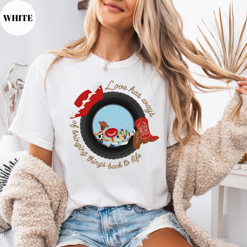 K&ouml;nnte beinhalten: Ein wei&szlig;es Kurzarm-T-Shirt mit einer Grafik eines schwarzen Reifens, der eine Szene mit Zeichentrickfiguren einrahmt. Das Design umfasst einen roten Cowboyhut, einen roten Stiefel und den braunen Schriftzug "Love has ways of bringing things back to life".