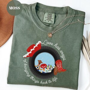 K&ouml;nnte beinhalten: Ein moosgr&uuml;nes Kurzarm-T-Shirt mit einer Grafik eines schwarzen Reifens, der eine Szene mit drei Zeichentrickfiguren einrahmt. Das Design enth&auml;lt einen roten Cowboyhut, einen roten Cowboystiefel und den Text Love has ways of bringing things back to life.