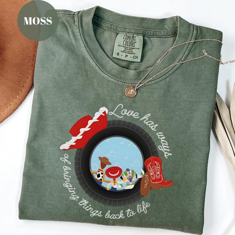 K&ouml;nnte beinhalten: Ein moosgr&uuml;nes Kurzarm-T-Shirt mit einer Grafik eines schwarzen Reifens, der eine Szene mit drei Zeichentrickfiguren einrahmt. Das Design enth&auml;lt einen roten Cowboyhut, einen roten Cowboystiefel und den Text Love has ways of bringing things back to life.