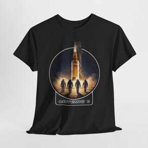 Könnte beinhalten: Schwarzes T-Shirt mit einem kreisförmigen Aufdruck, der einen Raketenstart und vier Astronauten zeigt. Die Rakete ist orange und weiß, vor einem dunklen, rauchigen Hintergrund. Der Text "ARTEMIS II" ist darunter in Weiß dargestellt.