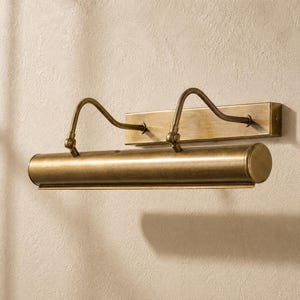 May include: A wall-mounted picture light fixture with an antique brass finish. The design features a rectangular backplate, two curved adjustable arms, and a long cylindrical light housing that directs illumination downward for artwork display.