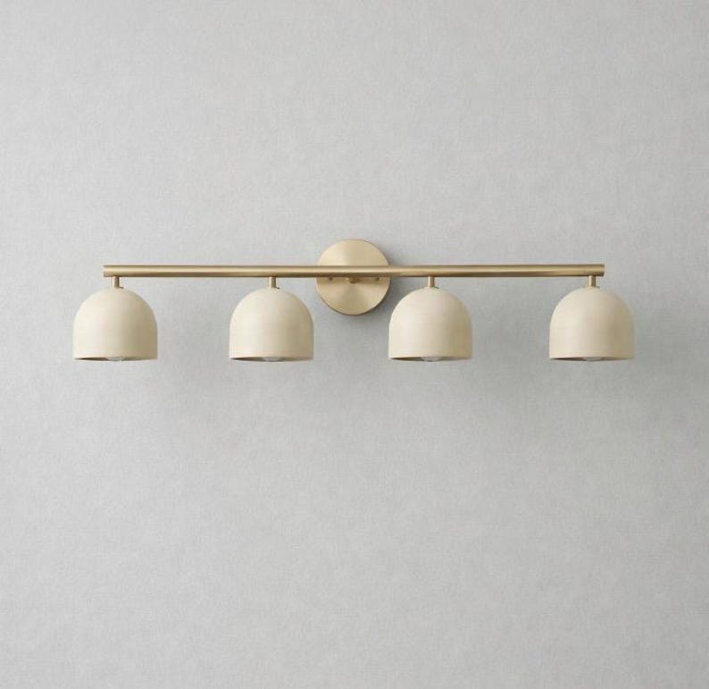 K&ouml;nnte beinhalten: Eine vierflammige Waschtischleuchte mit goldener Oberfl&auml;che. Die Leuchte hat vier cremefarbene, kuppelf&ouml;rmige Schirme. Das Licht ist an einer goldenen Stange und einer kreisf&ouml;rmigen Wandplatte befestigt.