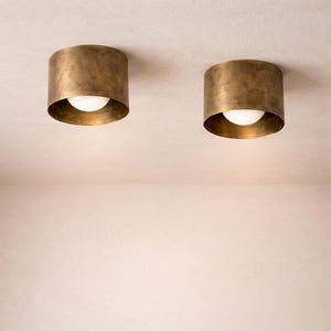 May include: Two cylindrical flush mount ceiling lights featuring a brushed antique brass finish. Each fixture has a wide, circular metal housing that encases a central, spherical white glass bulb, providing a soft, diffused light output.