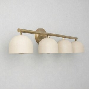 K&ouml;nnte beinhalten: Eine vierflammige Waschtischleuchte mit Messing-Finish. Sie hat vier cremefarbene, kuppelf&ouml;rmige Schirme. Das Licht ist an einer horizontalen Stange und einer kreisf&ouml;rmigen Basis befestigt.