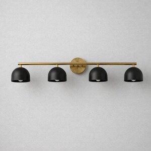 K&ouml;nnte beinhalten: Eine moderne Waschtischleuchte mit vier Lichtern. Sie verf&uuml;gt &uuml;ber vier schwarze, kuppelf&ouml;rmige Schirme, die an einer horizontalen Messingfarbenen Stange h&auml;ngen. Die Stange ist an einer kreisf&ouml;rmigen Wandhalterung in Messingfarbe befestigt.