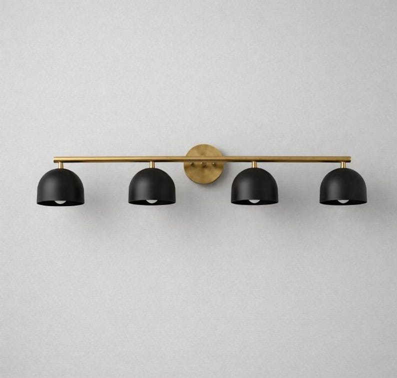 K&ouml;nnte beinhalten: Eine moderne Waschtischleuchte mit vier Lichtern. Sie verf&uuml;gt &uuml;ber vier schwarze, kuppelf&ouml;rmige Schirme, die an einer horizontalen Messingfarbenen Stange h&auml;ngen. Die Stange ist an einer kreisf&ouml;rmigen Wandhalterung in Messingfarbe befestigt.
