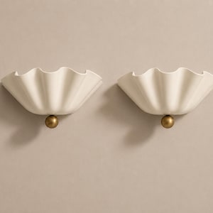 May include: A pair of wall sconces featuring off-white, ruffled, shell-shaped shades. Each sconce is accented with a single, centered, matte gold-toned sphere at the base, providing a clean, symmetrical design for wall-mounted lighting fixtures.